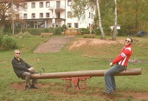 Zwei Männer auf einer Wippe, wobei der eine mit beiden Füßen auf dem Boden steht und den zweiten Mann den Boden nicht erreicht.